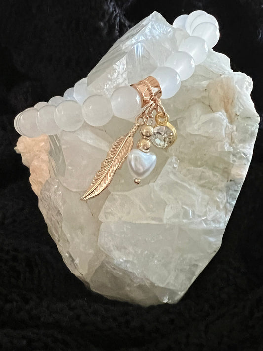 White beaded charm bracelet with gold accents atop a quartz crystal on black knit fabric.